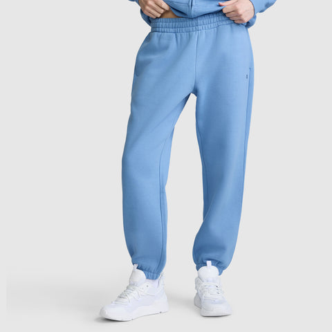 Wearing Champion Powerblend Oversized Sweatpants with C Logo in light blue, paired with white sneakers, she stands with hands in pockets against a plain light background.