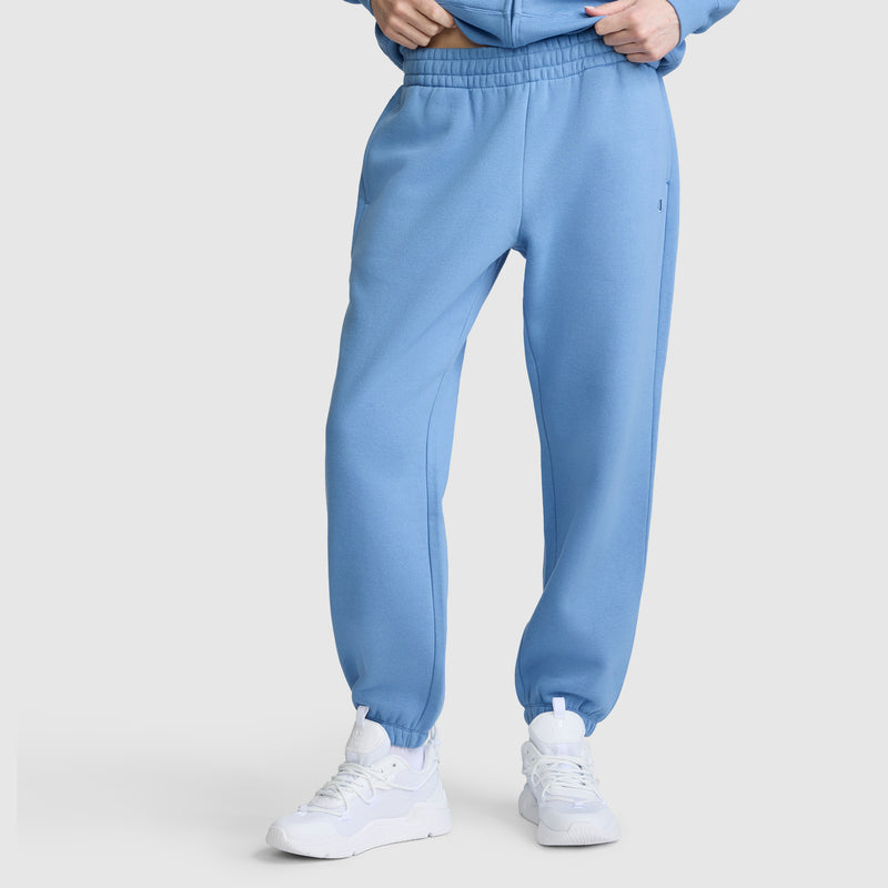 Wearing Champion Powerblend Oversized Sweatpants with C Logo in light blue, paired with white sneakers, she stands with hands in pockets against a plain light background.