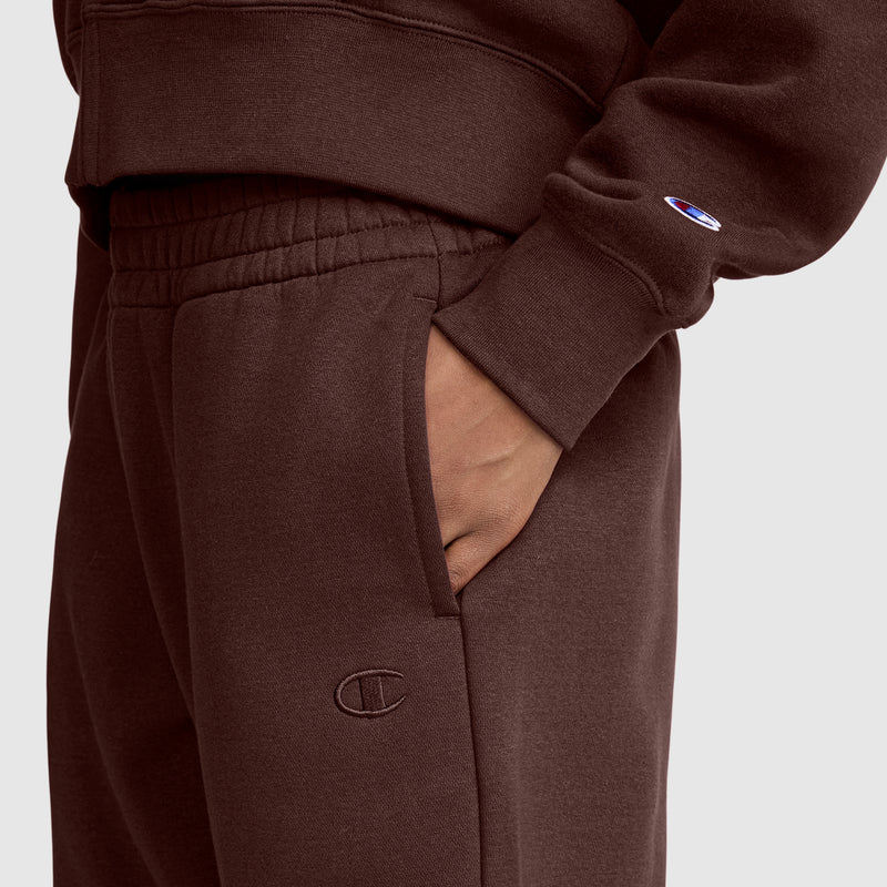 A person wears a brown Champion tracksuit. Their hand is in the pocket of the pants, which are the Champion Powerblend Oversized Sweatpants featuring the iconic C Logo. The sleeve displays a small embroidered Champion emblem. The oversized sweatpants give a relaxed look, set against a plain, light-colored background.
