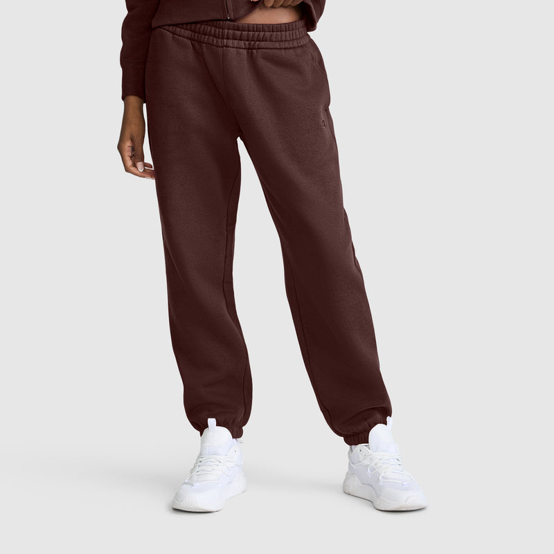 A person wearing maroon Champion Powerblend Oversized Sweatpants with a C Logo and white sneakers stands against a plain light gray background. Only the lower body and part of one arm are visible.
