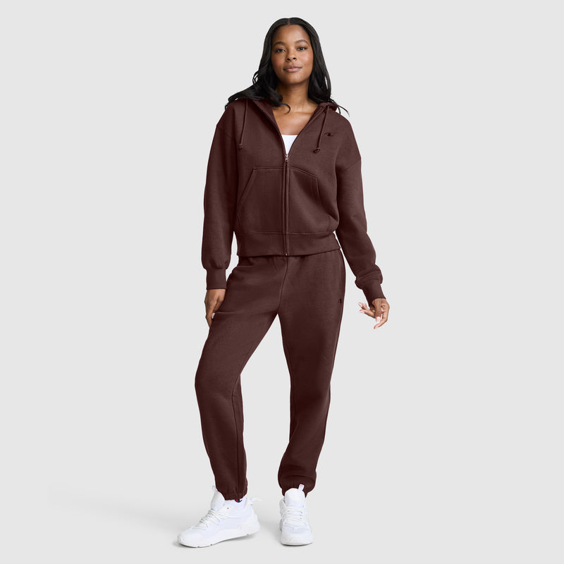 A woman stands against a plain light gray background wearing Champion’s Powerblend Oversized Sweatpants with the C Logo, paired with a matching maroon hoodie, a white top, and white sneakers.