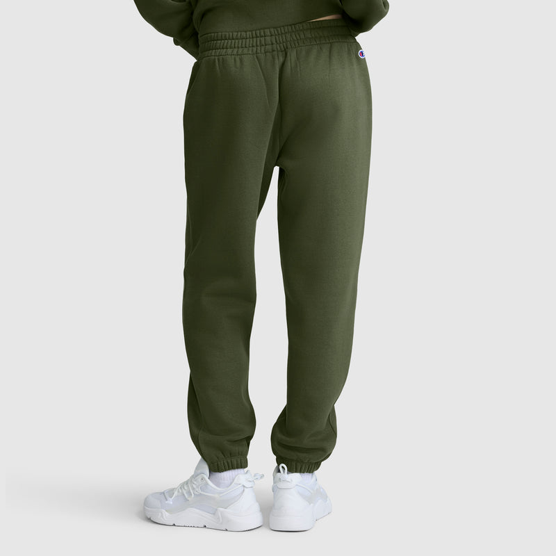 A person wearing Champion Powerblend Oversized Sweatpants with the C Logo in olive green and white sneakers stands facing away from the camera against a plain, light gray background.