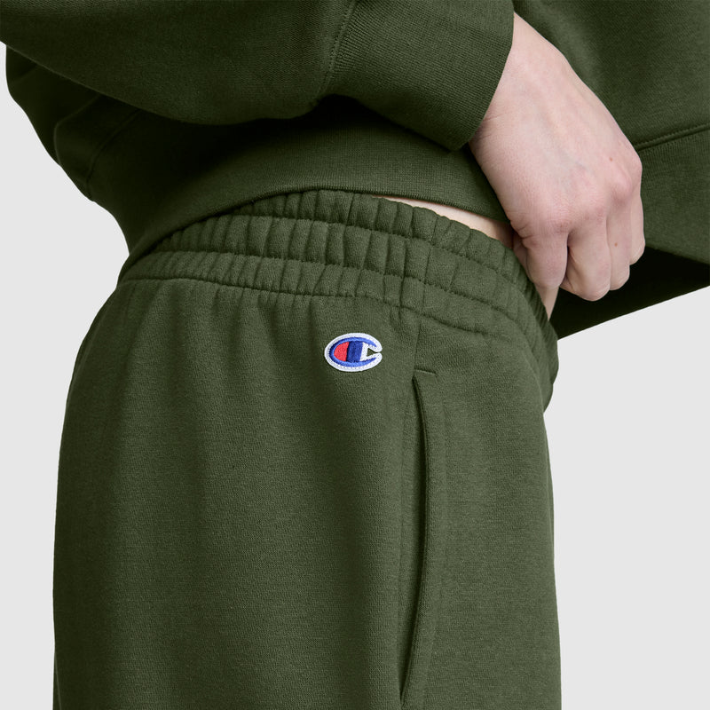 Wearing Champion’s Powerblend Oversized Sweatpants with the signature C logo on the pocket, a person adjusts the waistband for a perfect fit. The cozy fleece fabric ensures comfort while adding style to these olive green sweatpants.