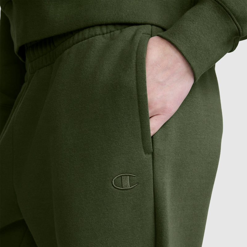 A person wearing dark green Champion Powerblend Oversized Sweatpants, C Logo, with a hand in the pocket. The oversized sweatpants feature the Champion C logo stitched near the pocket and are crafted from soft Powerblend fleece for comfort and style.