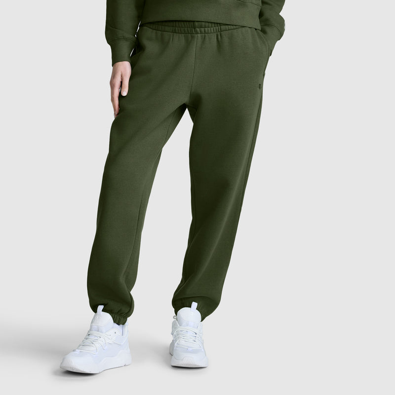 A person wearing Champion Powerblend Oversized Sweatpants, C Logo in olive green and white sneakers stands against a plain light gray background, with only the lower half of their body visible.