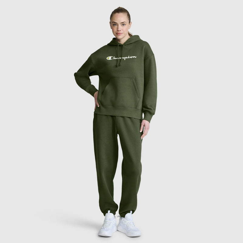 A woman stands facing forward against a plain light gray background, wearing a dark green Champion hoodie paired with Champion’s Powerblend Oversized Sweatpants featuring the C Logo, both crafted from cozy Powerblend fleece, and finished with white sneakers.