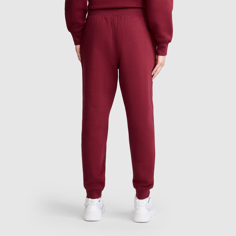 A person wearing maroon Champion Powerblend Joggers, C Logo, with a matching sweatshirt is shown from the back standing against a plain light gray background. They are also wearing white sneakers.