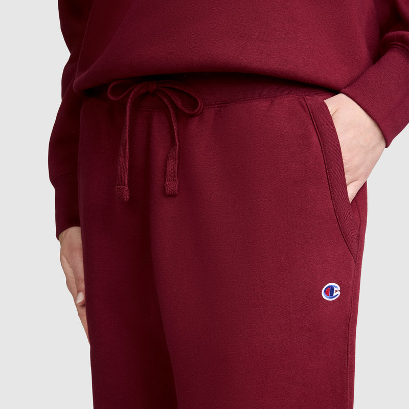 A person is wearing maroon Champion Powerblend Joggers, C Logo, paired with a matching sweatshirt and has one hand in their pocket. The Powerblend fleece joggers feature a small embroidered Champion C logo near the pocket. The background is plain and light-colored.