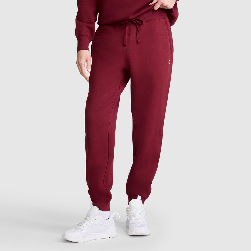 A person is wearing maroon Champion Powerblend Joggers, C Logo, paired with a matching sweatshirt and white sneakers. They are standing with one hand in their pocket against a plain, light-colored background.