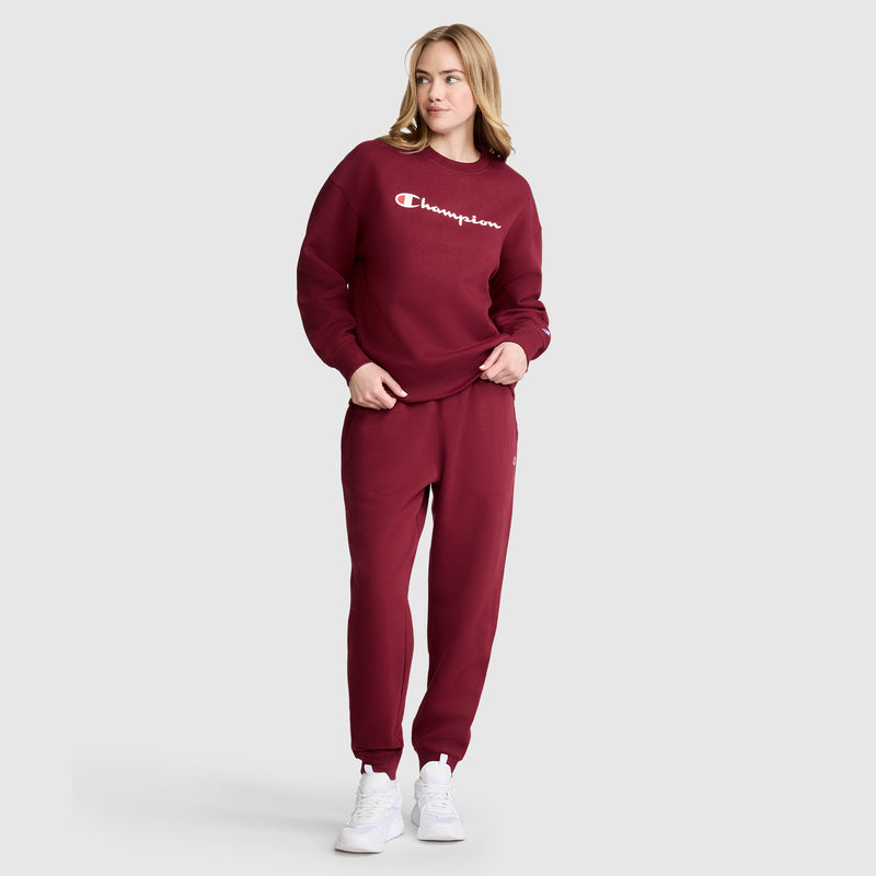 A woman wearing a maroon Champion sweatshirt and matching Powerblend Joggers, C Logo by Champion stands with one hand in her pocket, looking to the side against a plain light gray background.