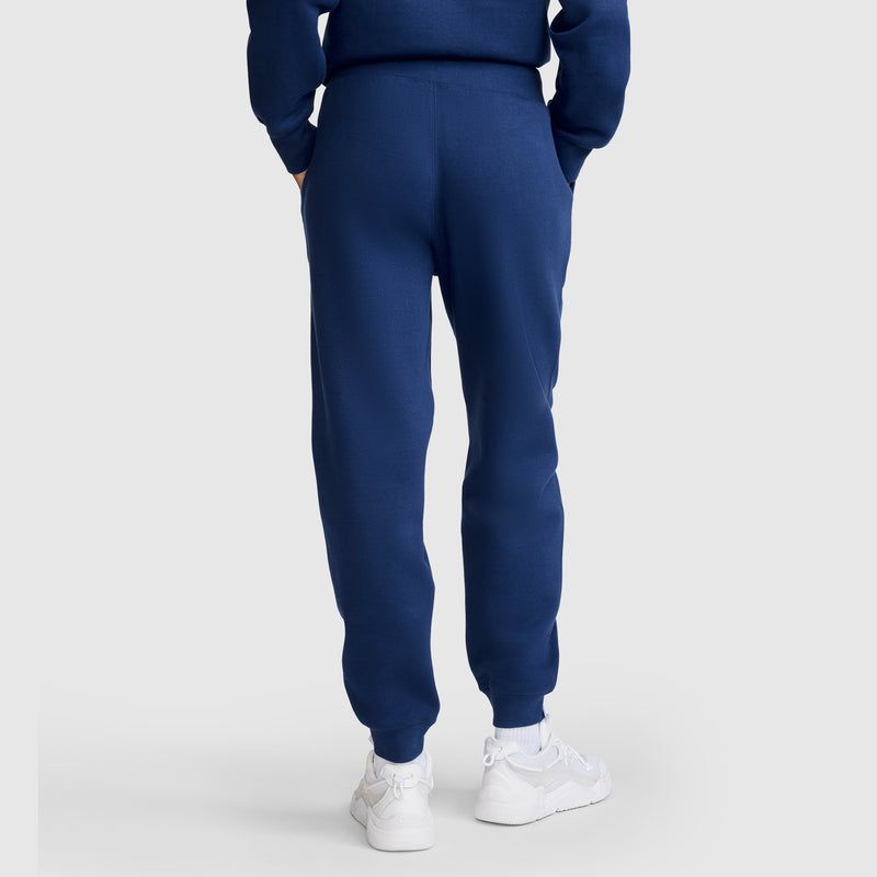 A person stands with hands in pockets, facing away from the camera against a plain light background, wearing blue Champion Powerblend Joggers with a C Logo and a matching blue sweatshirt, paired with white sneakers.