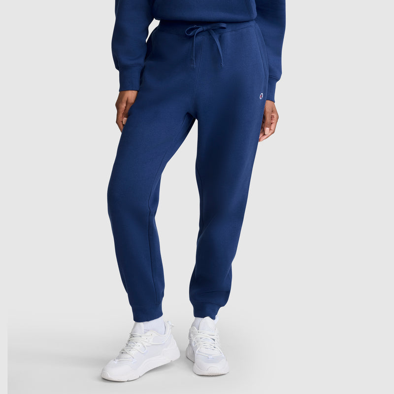 Wearing Champion’s Powerblend Joggers with the C Logo in blue, paired with a matching blue sweatshirt and white sneakers, this person stands against a plain light gray background.