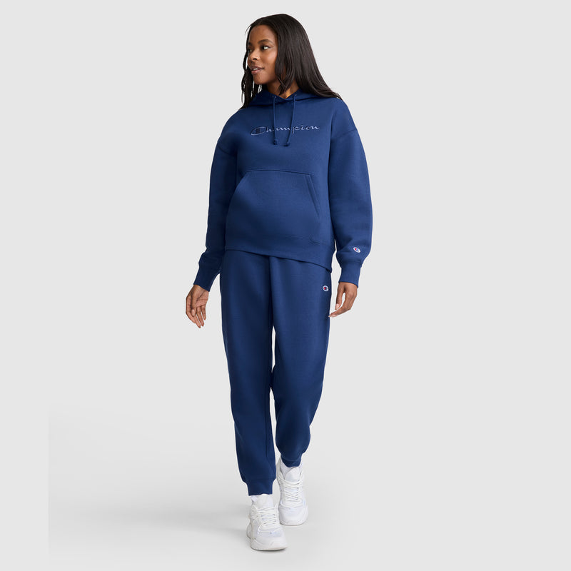 A woman wearing Champion Powerblend Joggers with the C Logo and a coordinating blue hoodie, paired with white sneakers, walks on a plain light background, looking to her left and smiling slightly.