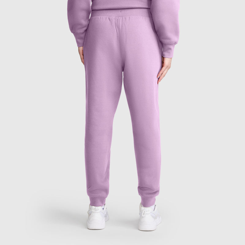 A person stands facing away, wearing Champion Powerblend Joggers with a C Logo in light purple matched with a coordinating sweatshirt and white sneakers. The background is plain and light-colored.