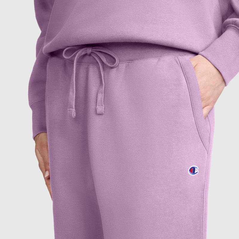 A person wearing Champion Powerblend Joggers, C Logo in lavender, with one hand in their pocket. The Champion logo patch is visible near the pocket, highlighting these stylish fleece jogger pants. A matching sweatshirt from Champion is partially visible.