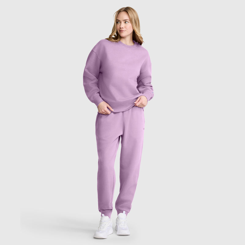 A woman stands wearing Champion Powerblend Joggers with a C Logo in light purple, paired with a matching sweatshirt and white sneakers, posing casually against a plain light background.