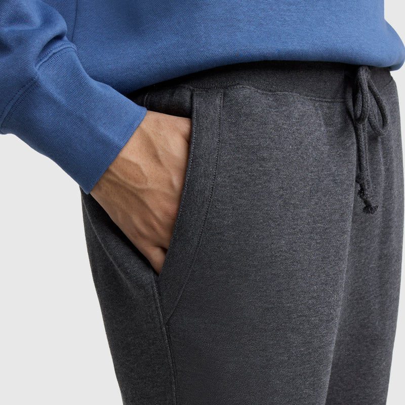 Someone is shown from the waist down against a plain background, wearing Champion Powerblend Straight Hem Sweatpants in dark gray with their right hand in their pocket and a blue sweatshirt.