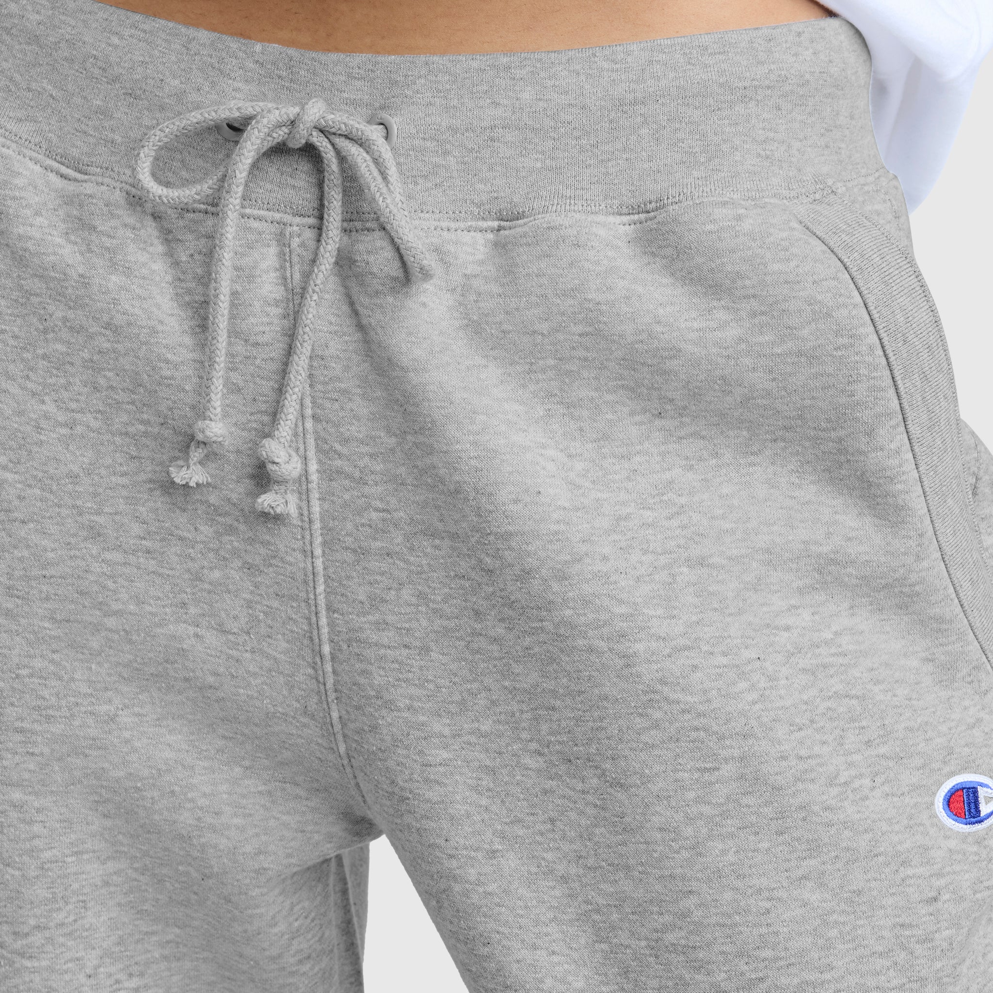 Close-up of a person wearing Champion Powerblend Straight Hem Sweatpants in light gray midweight fleece, featuring the small blue, red, and white logo on the left thigh. The white shirt is tucked in slightly.