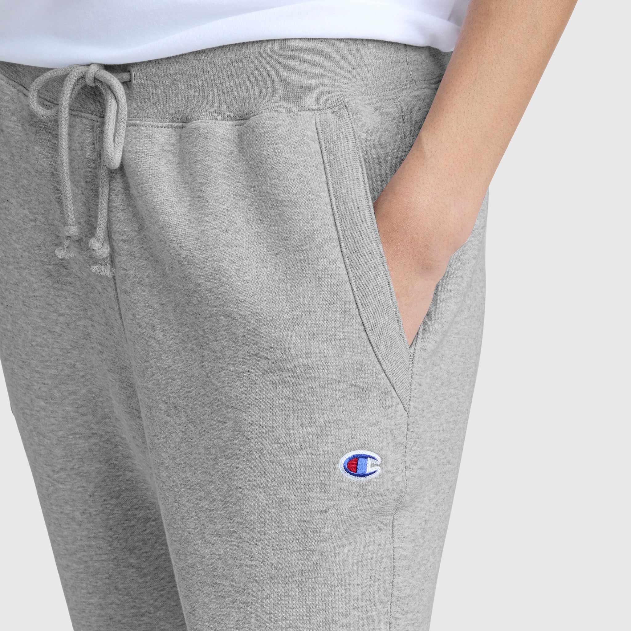 Someone wearing Champion Powerblend Straight Hem Sweatpants with a drawstring and the Champion logo on the pocket stands with one hand in their pocket against a plain background.