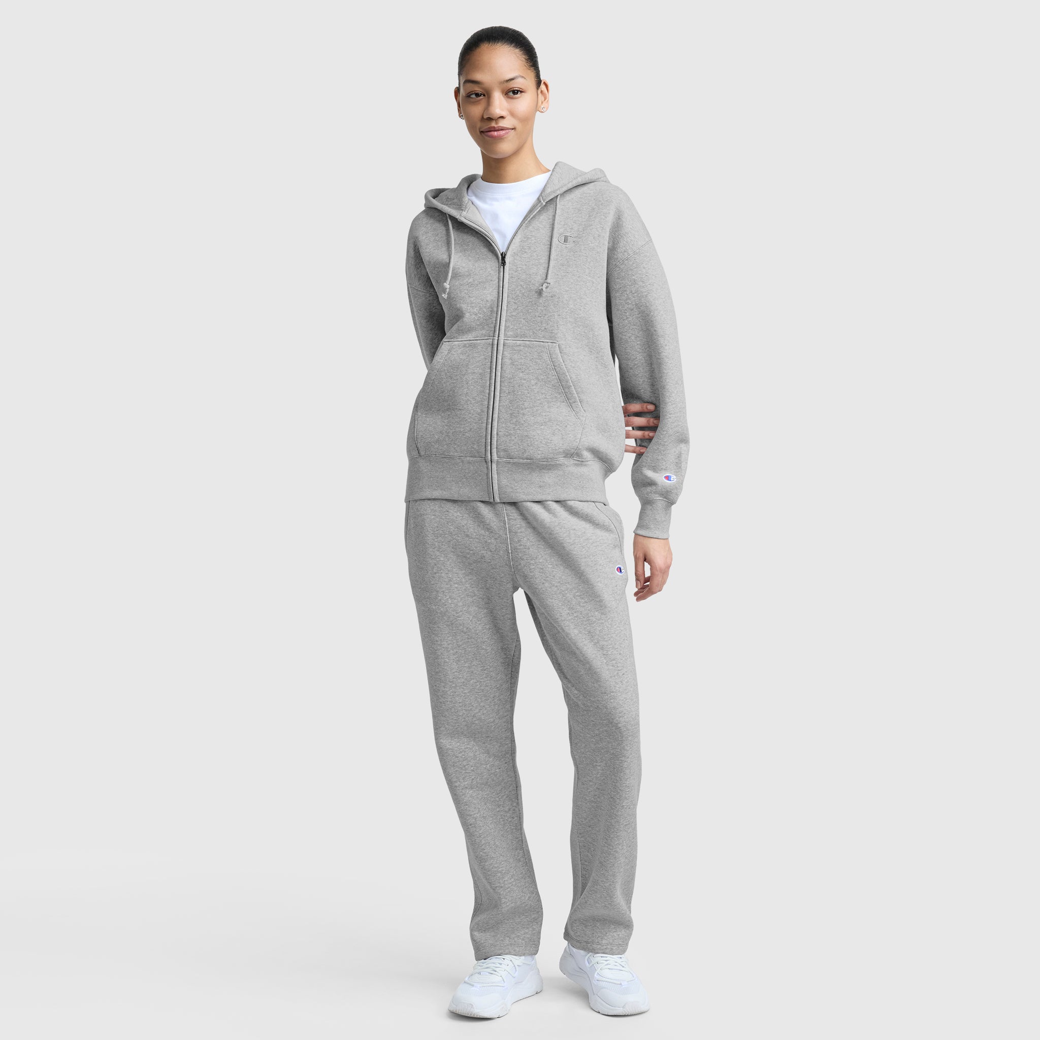 A person stands against a plain background wearing a light gray zip-up hoodie, Champion Powerblend Straight Hem Sweatpants, white sneakers, and a white t-shirt. They have a relaxed posture and a neutral expression.