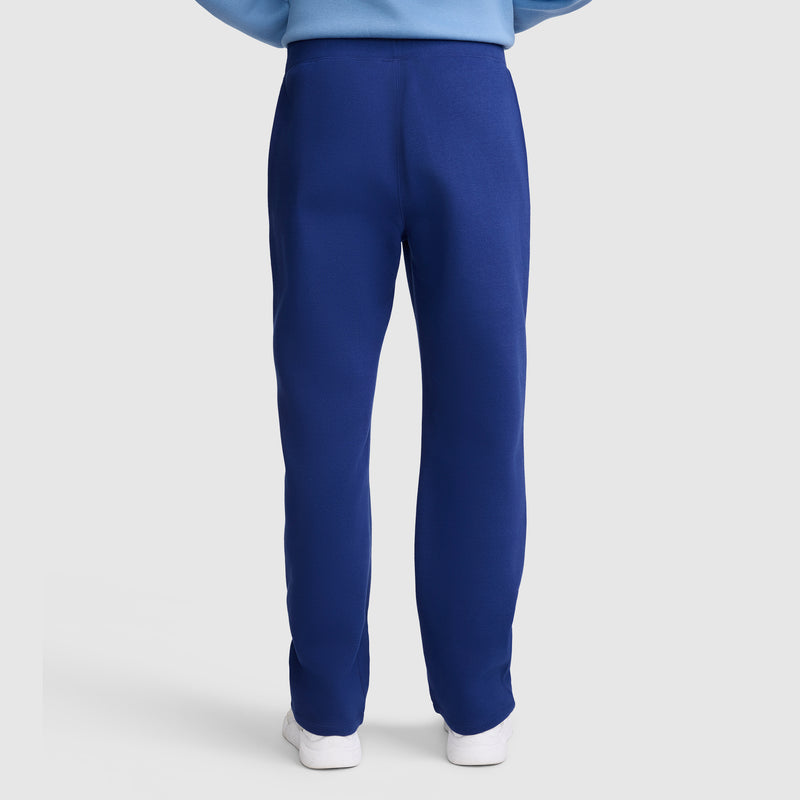 A person stands with their back to the camera, wearing Champion Powerblend Straight Hem Sweatpants in bright blue and a light blue sweatshirt. The full-length, slim-fit sweatpants are crafted from midweight fleece for comfort and style, paired with white shoes against a plain, light-colored background.