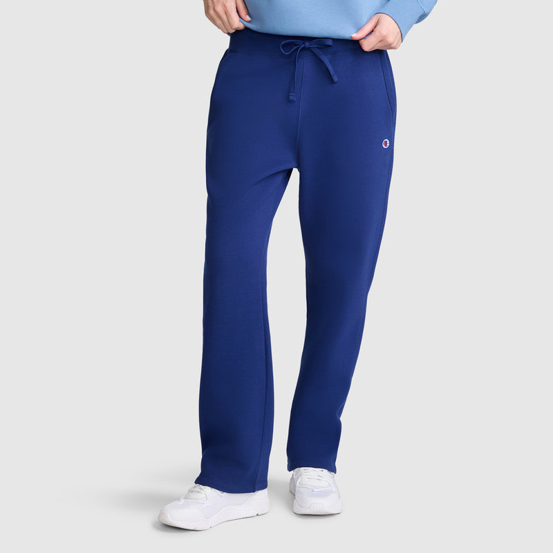A person wearing Champion Powerblend Straight Hem Sweatpants in royal blue—crafted from midweight fleece with a slim fit, drawstring waist, and a small logo on the left thigh—paired with white sneakers and a light blue top, stands against a plain light background.