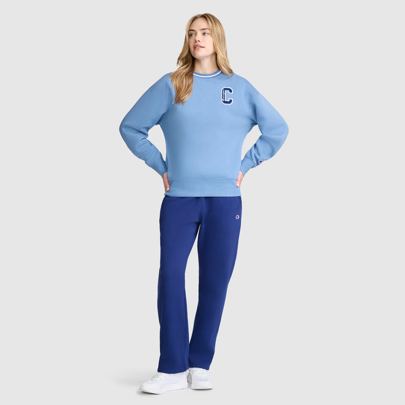 A woman stands with hands on hips, wearing a light blue, midweight fleece sweatshirt with a C patch, paired with slim fit dark blue Champion Powerblend Straight Hem Sweatpants and white sneakers against a plain light gray background.