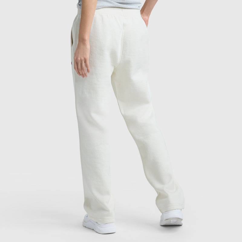 A person stands on a white background wearing Champion Powerblend Straight Hem Sweatpants in off-white with white sneakers, one hand in their pocket and their back turned to the camera.