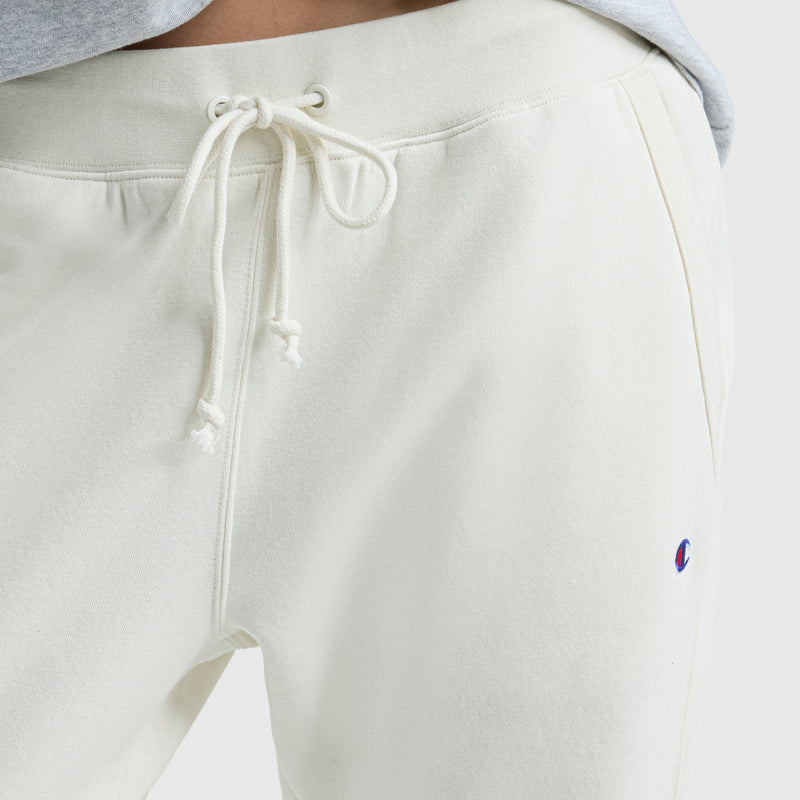 Close-up of a person wearing Champion Powerblend Straight Hem Sweatpants in off-white midweight fleece, with a drawstring waist and small blue and red embroidered logo on the left leg; gray shirt partially visible above waistband.