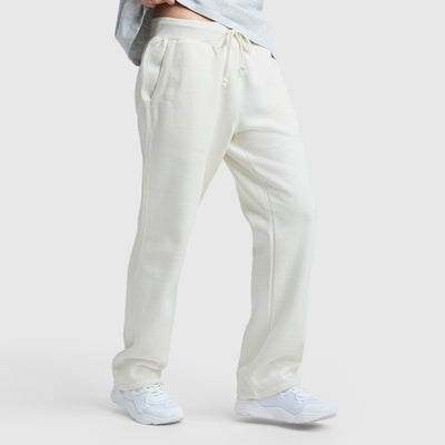 A person wears cream-colored Champion Powerblend Straight Hem Sweatpants made from midweight fleece and white sneakers, standing against a plain light background. The shot focuses on the lower body with hands in pockets and a straight hem finish.