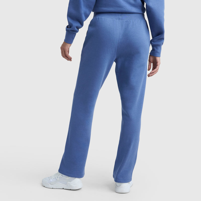 A person stands with their back to the camera, wearing blue Champion Powerblend Straight Hem Sweatpants and a matching blue midweight fleece sweatshirt. Both have straight hems and are paired with white sneakers against a plain white background.