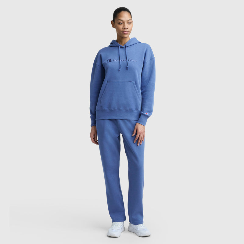 A woman wears the Champion Powerblend Straight Hem Sweatpants with a matching blue hoodie featuring a front pocket and chest logo. The set is made from midweight fleece and paired with white sneakers against a plain white background.
