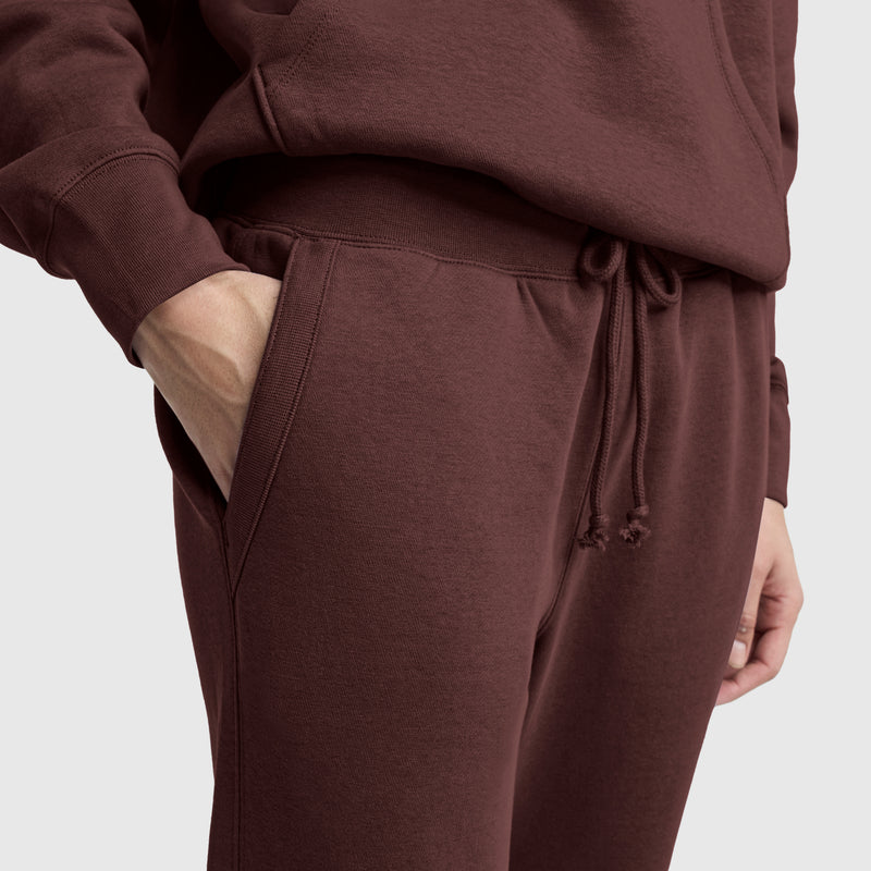 Someone wearing a matching maroon hoodie and Champion Powerblend Straight Hem Sweatpants, with one hand in a pocket, stands against a plain light background. Only the torso and upper legs are visible.