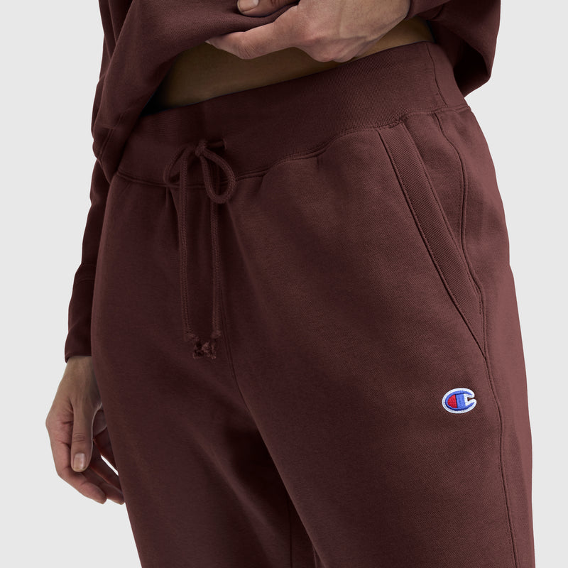 A person wearing Champion Powerblend Straight Hem Sweatpants in a slim fit maroon style, crafted from midweight fleece with a drawstring and featuring the iconic Champion logo, stands against a plain background with one hand in their pocket and the other resting on their hip.