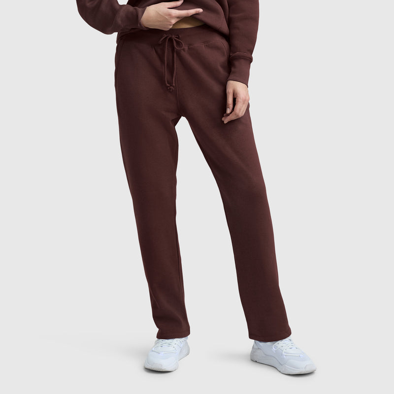 A person wearing Champion Powerblend Straight Hem Sweatpants in brown, a matching brown sweatshirt, and white sneakers stands against a plain light gray background.