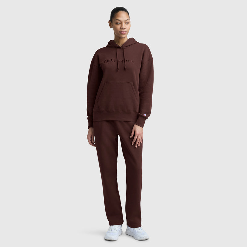 A woman wearing a matching brown slim fit hoodie and Champion Powerblend Straight Hem Sweatpants with white sneakers stands on a plain light gray background, looking slightly to the side with one hand by her side and the other resting on her leg.