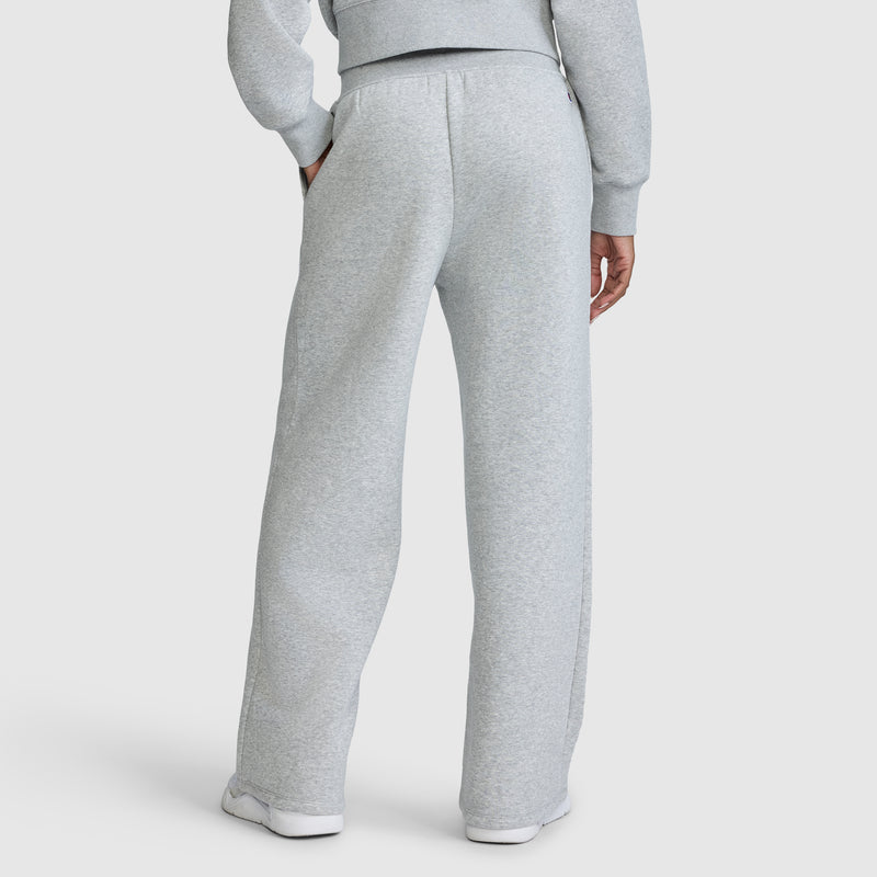 A person is seen from behind standing against a plain white background, wearing Champion’s Wide Leg Fleece Sweatpants featuring the C Logo along with a matching light gray sweatshirt. One hand is in their pocket and they are wearing white shoes.