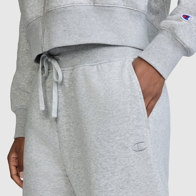 A person is wearing Champion’s Wide Leg Fleece Sweatpants featuring the signature C Logo, with their hand in the pocket; a matching sweatshirt from Champion is partially visible. Both pieces display the recognizable Champion branding. Set against a plain, light-colored background, these sweatpants offer a cozy and comfortable style statement.
