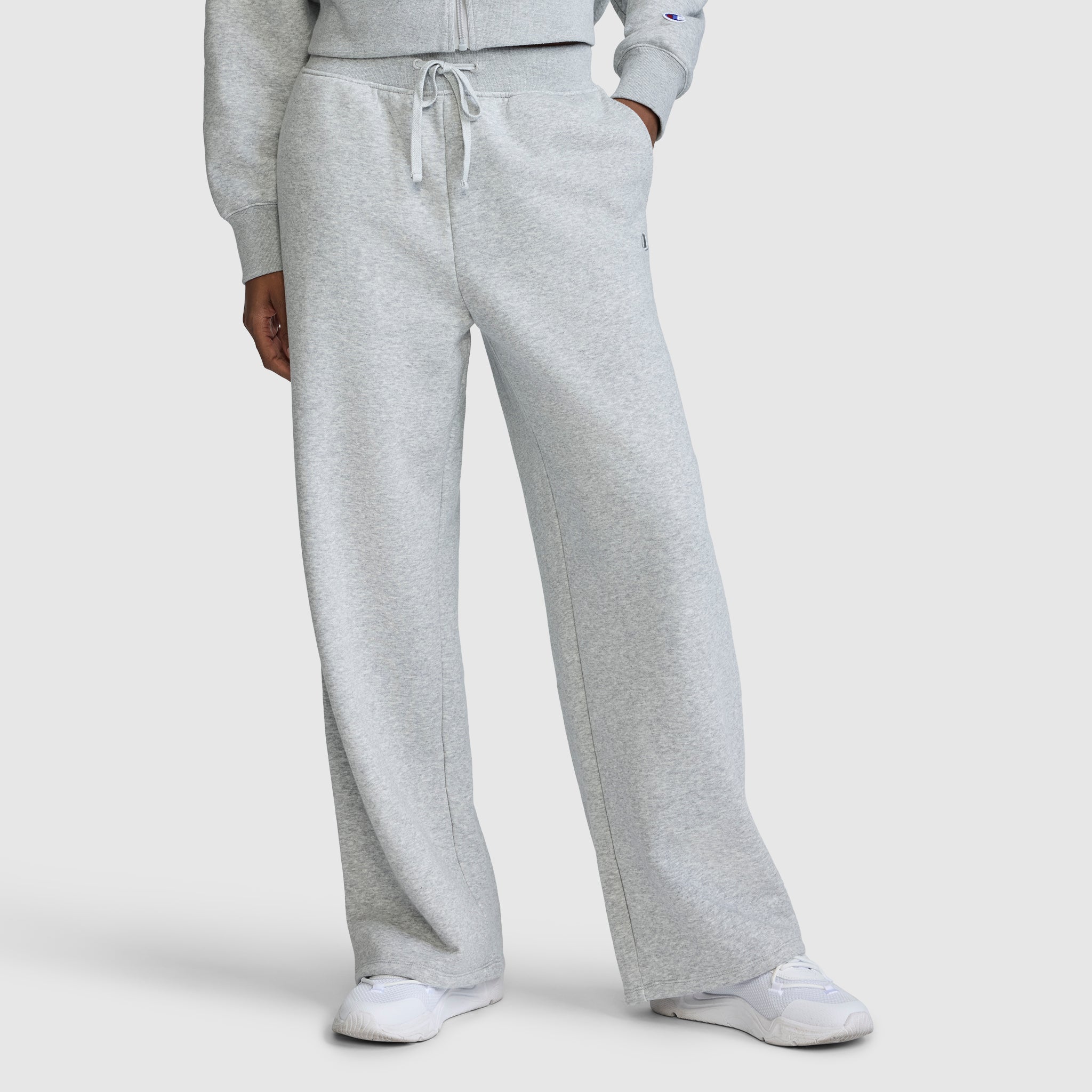 A person is wearing Champion's Wide Leg Fleece Sweatpants with the C Logo in light gray, paired with a matching sweatshirt and white sneakers. The photo is cropped from the shoulders down to showcase the relaxed fit and comfortable style of these sweatpants.