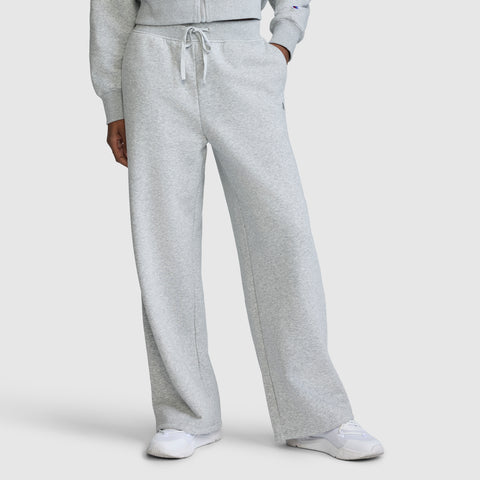A person is wearing Champion's Wide Leg Fleece Sweatpants with the C Logo in light gray, paired with a matching sweatshirt and white sneakers. The photo is cropped from the shoulders down to showcase the relaxed fit and comfortable style of these sweatpants.