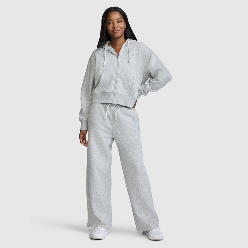 A woman stands confidently in a light gray zip-up hoodie and matching Champion Wide Leg Fleece Sweatpants, C Logo, paired with white sneakers against a simple light background—a sleek ensemble ideal for modern women's loungewear.