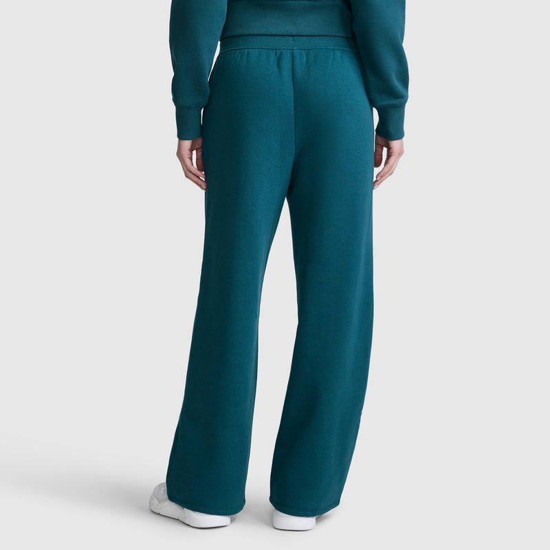 A person is seen from behind wearing Champion's Wide Leg Fleece Sweatpants with a C Logo in teal, paired with a matching long-sleeve top and white sneakers. The background is plain and light-colored.
