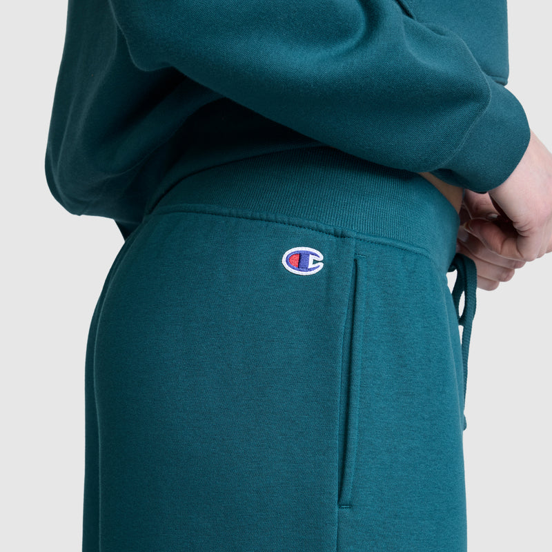 A person is wearing Champion’s Wide Leg Fleece Sweatpants, featuring the signature C logo near the pocket. The teal women’s sweatpants look cozy and are paired with a matching sweatshirt. The person’s hand rests near their hip against a plain light gray background.
