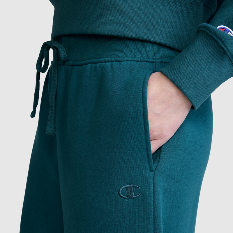 A close-up shows a person wearing Champion’s Wide Leg Fleece Sweatpants, C Logo, in teal, paired with a matching sweatshirt. One hand is tucked into the pocket of the cozy wide leg fleece sweatpants, which feature the iconic Champion logo on both the pants and sleeve for a signature finishing touch.