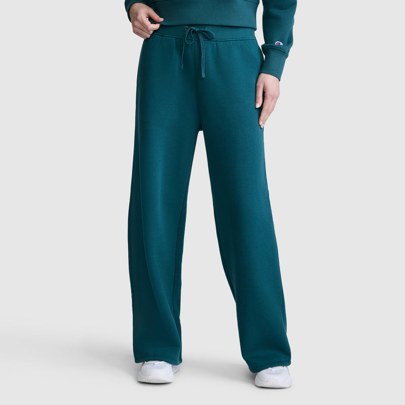 A person is shown from the waist down, standing against a plain white background, wearing Champion Wide Leg Fleece Sweatpants with the C Logo paired with a matching teal sweatshirt and white sneakers.