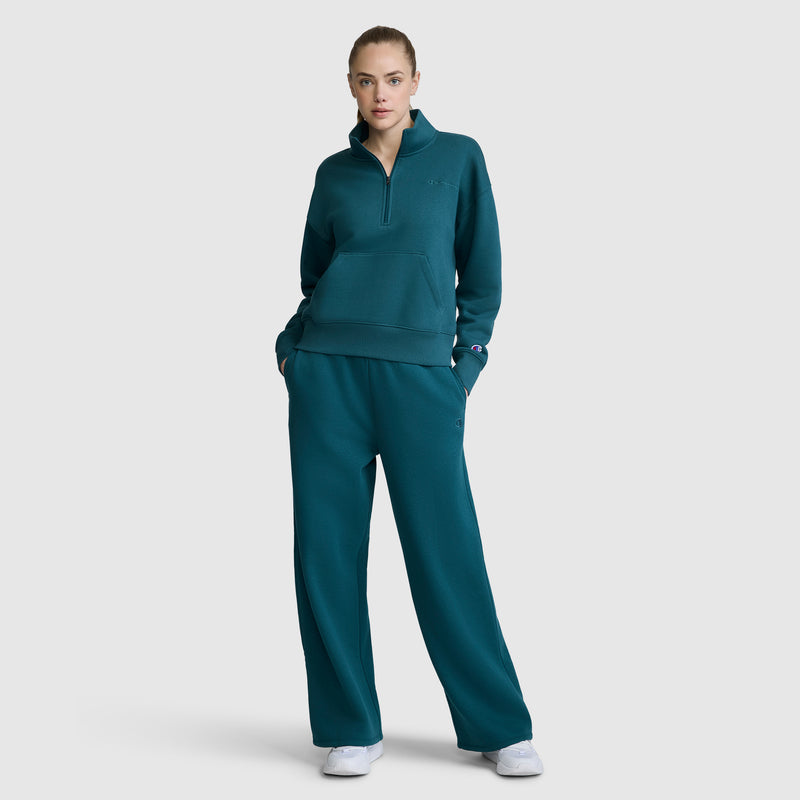 A person stands facing forward with hands in their pockets, wearing Champion’s Wide Leg Fleece Sweatpants with the C Logo in teal, paired with a matching pullover sweatshirt and white sneakers, against a plain light gray background.