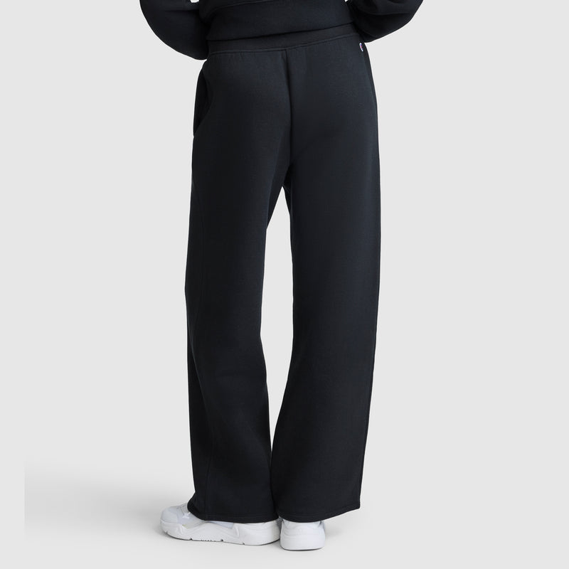 A person is seen from behind, standing against a light gray background, wearing Champion Wide Leg Fleece Sweatpants, C Logo in black paired with white sneakers. The full length of the women's sweatpants is visible in the photo.