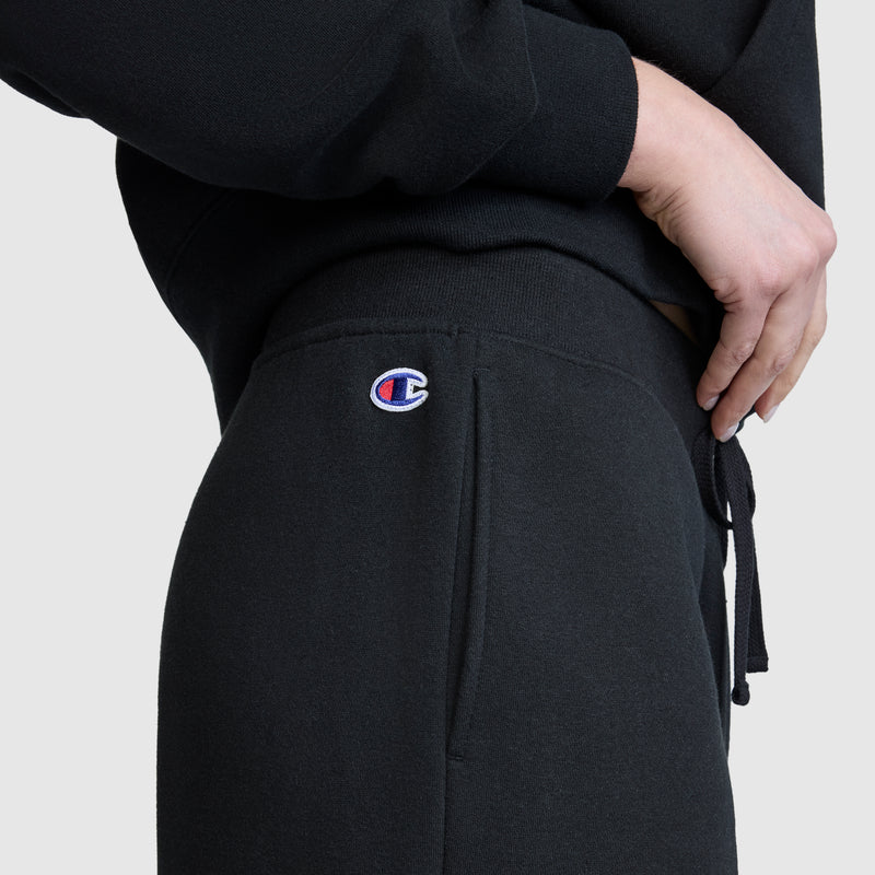 A close-up shows a person wearing Champion's Wide Leg Fleece Sweatpants with the signature C logo near the pocket. The individual, whose hand rests on their hip, is also dressed in a black sweatshirt. These comfortable women's sweatpants are ideal for lounging or everyday wear, set against a plain, light-colored background.