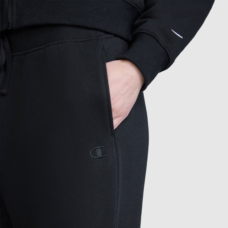 A person wearing Champion Wide Leg Fleece Sweatpants, C Logo in black and a black hoodie has their hand in their pocket. The Champion logo is visible on the women's sweatpants. The background is plain light gray.