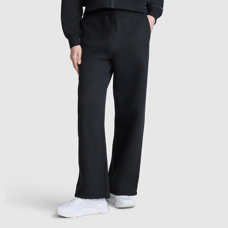 A person wearing black Champion Wide Leg Fleece Sweatpants with a C Logo, a matching black sweatshirt, and white sneakers stands against a plain light gray background.